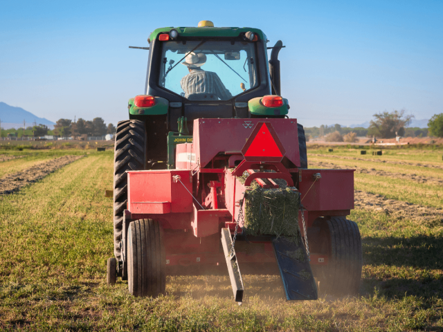 A farmer drives a tractor and hay baler through a field, producing rectangular hay bales.