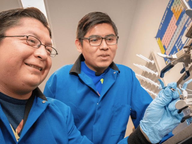 Two researchers in blue lab coats examine a sample secured in laboratory equipment.