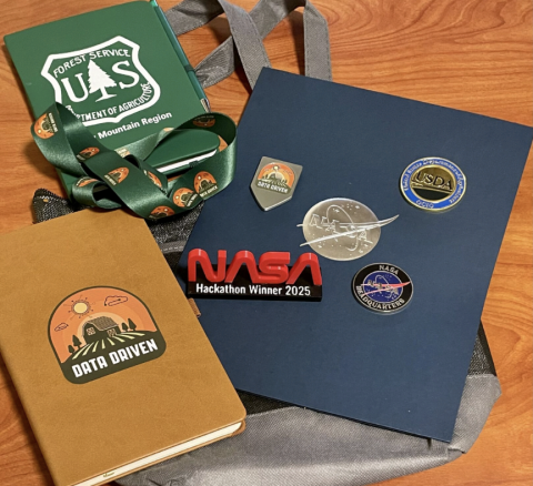 Picture shows the available competition student prizes, including USDA FS and Data-Driven notebooks, lanyards, challenge coins from USDA and NASA, and a NASA Hackathon Winner 2025 award