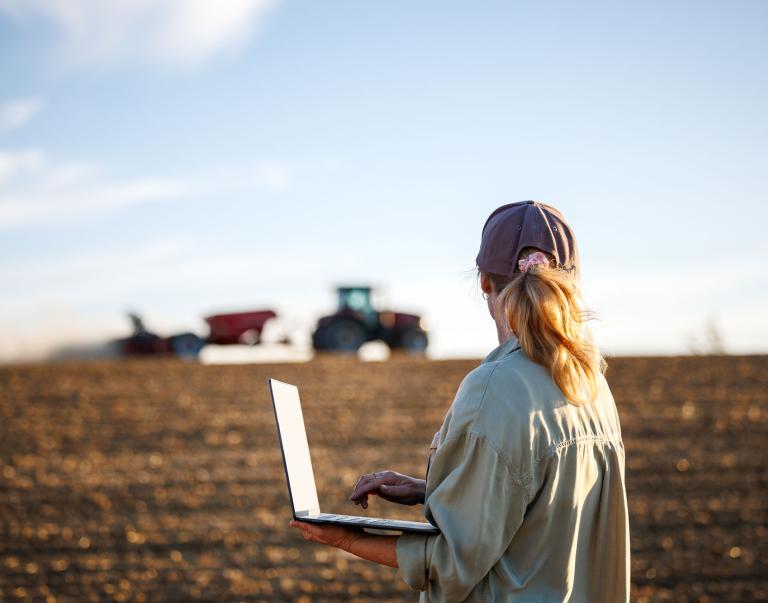 Woman farmer with laptop analyzing agricultural data in front of tractor and seeding machine on cultivated field
