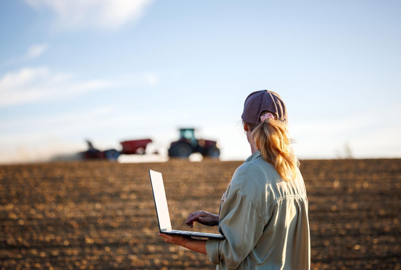 Woman farmer with laptop analyzing agricultural data in front of tractor and seeding machine on cultivated field